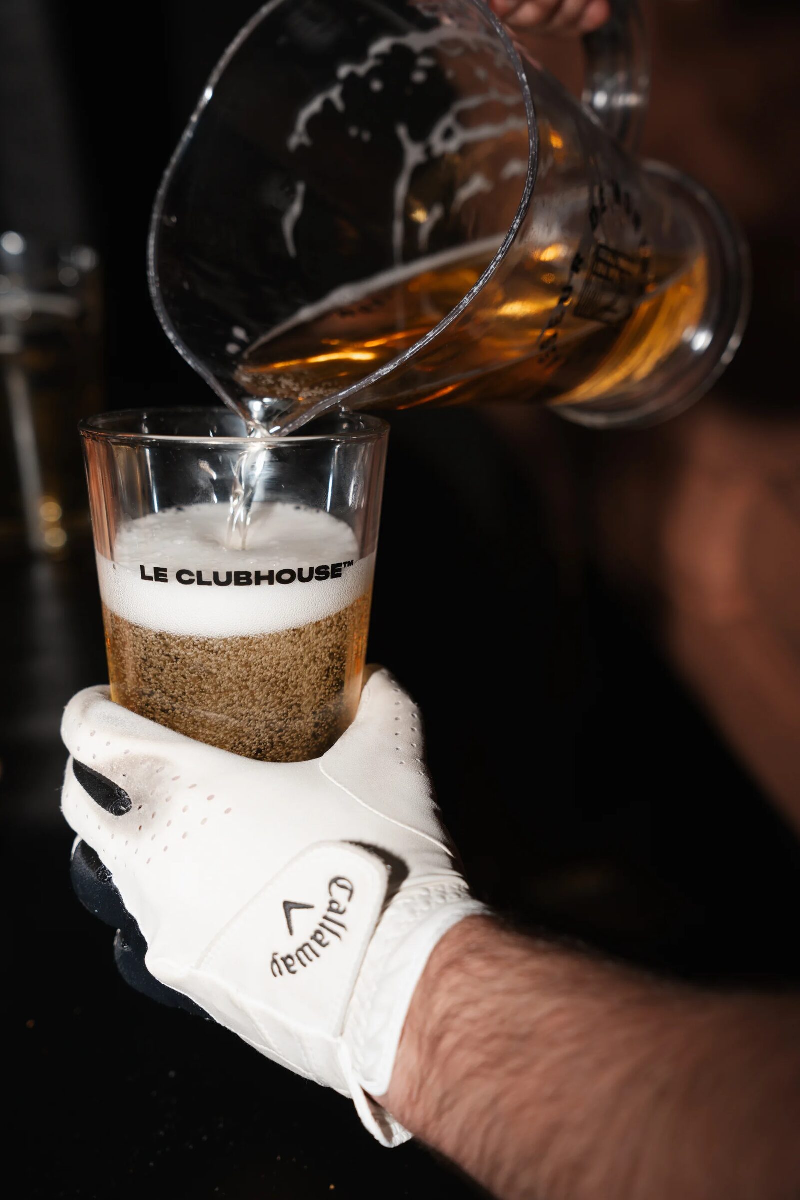Un pichet de bière versé dans un verre Le Clubhouse, tenu par une main gantée