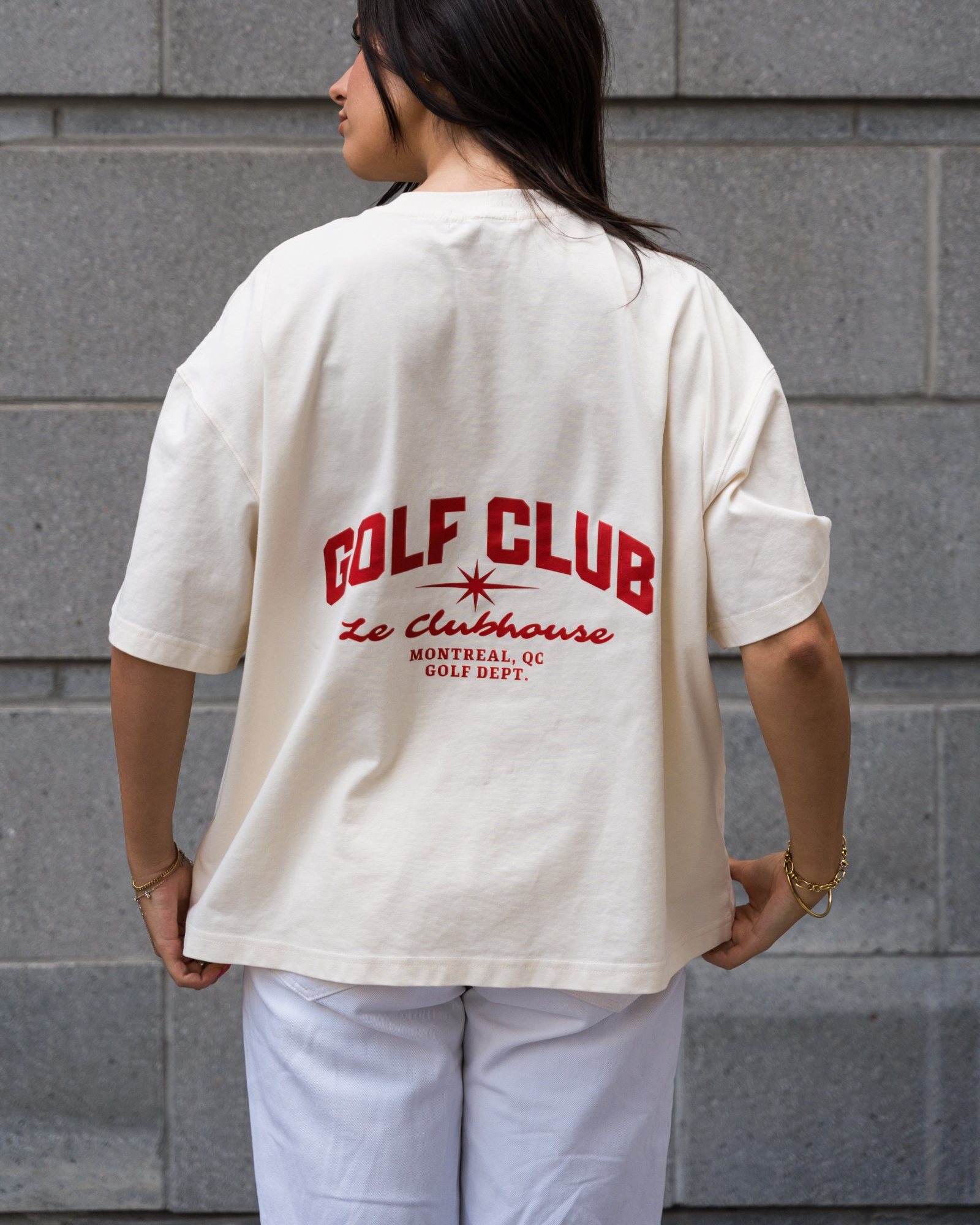Une cliente de dos qui porte le t-shirt crème GOLF CLUB Le Clubhouse, lettrage rouge, devant un mur de pierre grise