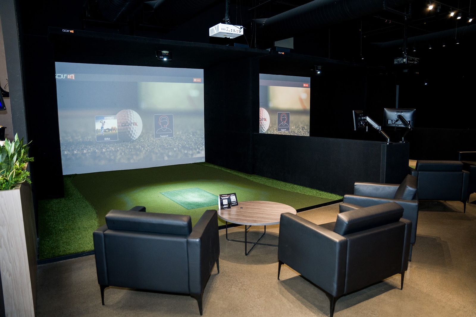 Une baie avec deux fauteuils carrés en cuir noir et une table ronde devant l'écran du simulateur de golf