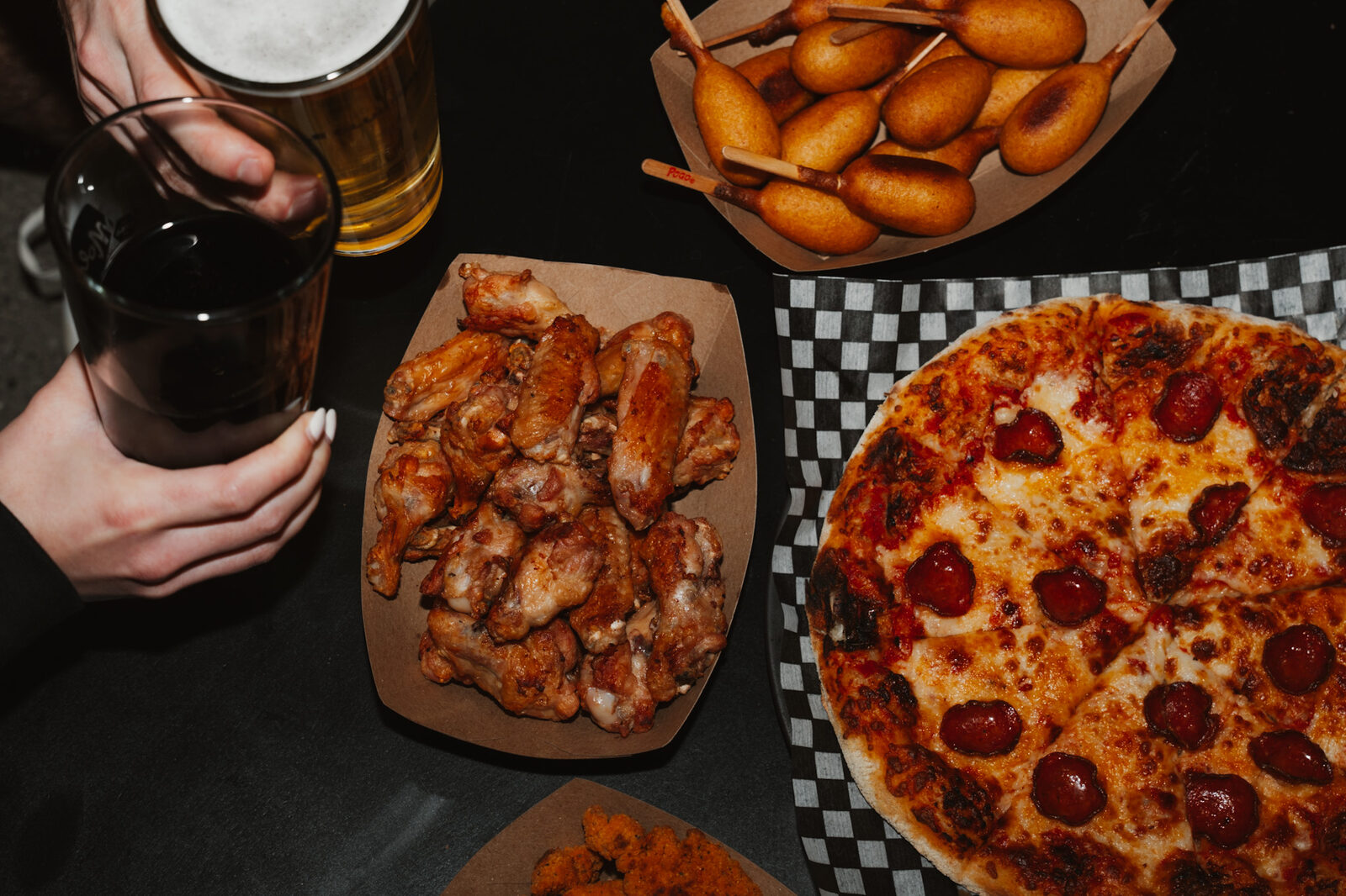 Vue de haut d'une table du Clubhouse : une pizza pepperoni, des ailes de poulet, des mini pogos et deux verres de bière tenus à la main